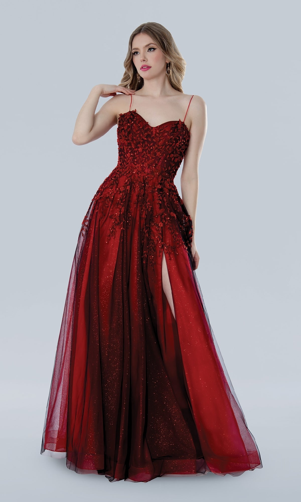 A woman in the Stella Couture 25109 long prom dress—sleeveless, floor-length red lace with floral embroidery and a thigh-high slit—stands against a plain light background.